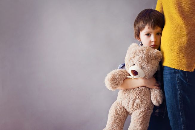 Image of small child with teddy bear holding onto mom's leg