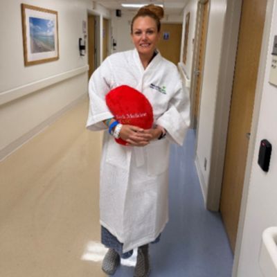 Tiffaney in a hospital gown stands in a hospital hallway holding a red heart-shaped pillow that reads “You Matter.”